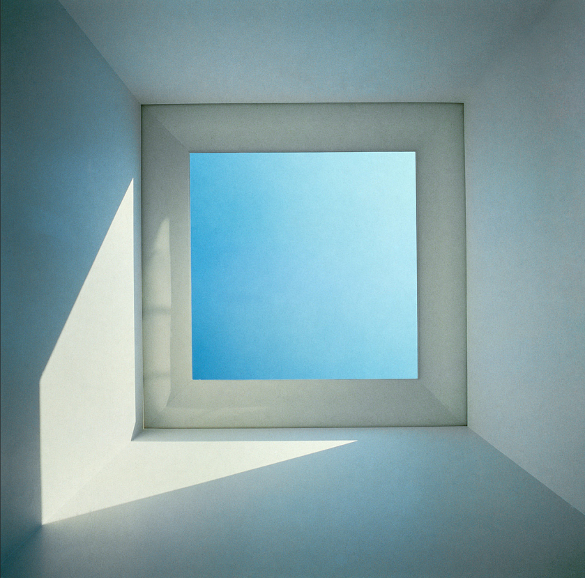 Picture of the ceiling of a white room. The ceiling is square and in the middle there is a square window/hole. Trough it you can view a blue sky. It is the artwork skyspaces from James Turrell. Where he plays with light.